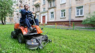 Рабочий косит траву газонокосилкой во дворе многоквартирного дома.