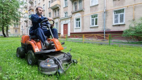 Рабочий косит траву газонокосилкой во дворе многоквартирного дома.