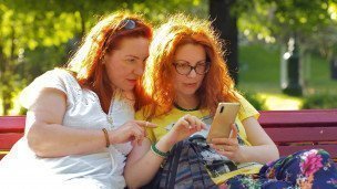 Две девушки сидят на скамейке в парке и смотрят в смартфон