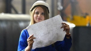 Девушка в каске и рабочей форме держит в руках чертеж и смотрит в даль  