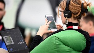 Девушка в наушниках сидит спиной к зрителю на коленях у нее ноутбук а в руках смартфон