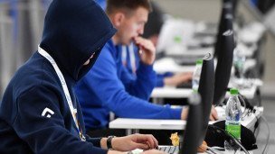 Человек в капюшоне сидит за компьютером за ним в ряд еще несколько людей за компьютерами 