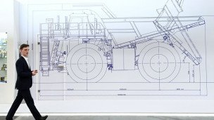 Посетитель в деловом костюме на международной промышленной выставке идет мимо чертежа экскаватора 