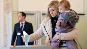 Женщина с ребенком на руках опускает бюллетень в урну для голосования